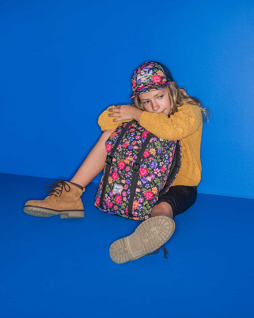 A child is sitting on the ground with a Retreat Backpack Youth in Garden Floral