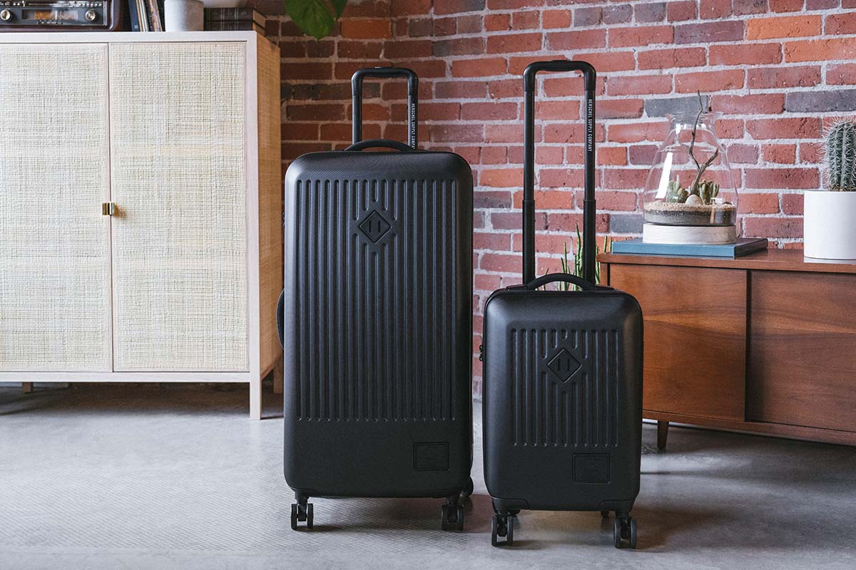 Un Trade Luggage Large et un Trade Luggage Carry-On noirs à côté l’un de l’autre dans un appartement en briques apparentes
