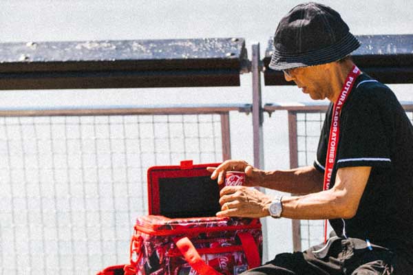 A person sitting on a bench on a pier wearing the Futura Laboratories x Coca-Cola® Lanyard while grabbing a Coke outta the Insulated 12 Pack Cooler.