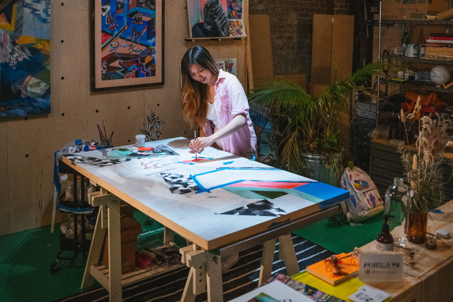 A person in their art studio painting a large canvas on the table with their artwork hanging behind them and their Heavyweight Canvas Herschel Heritage Backpack on the ground behind them with paint all over it.