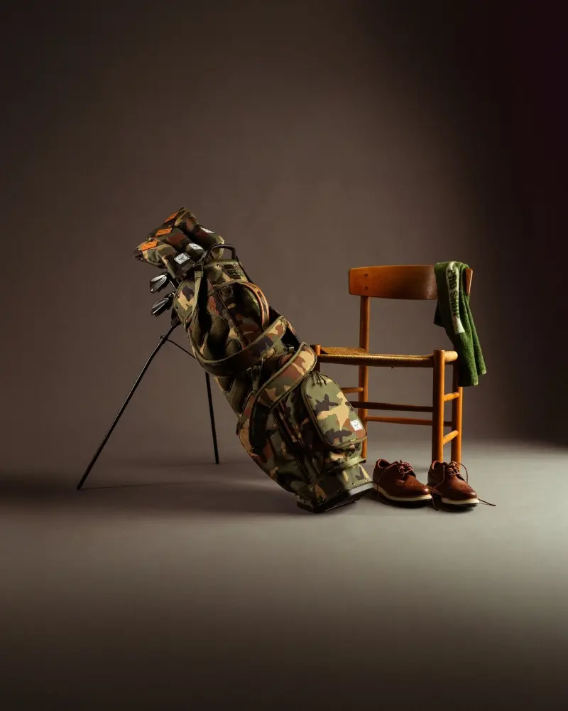 A woodland camo herschel supply golf bag (watt stand bag) sits on its pop out legs against a grey gradient studio background 